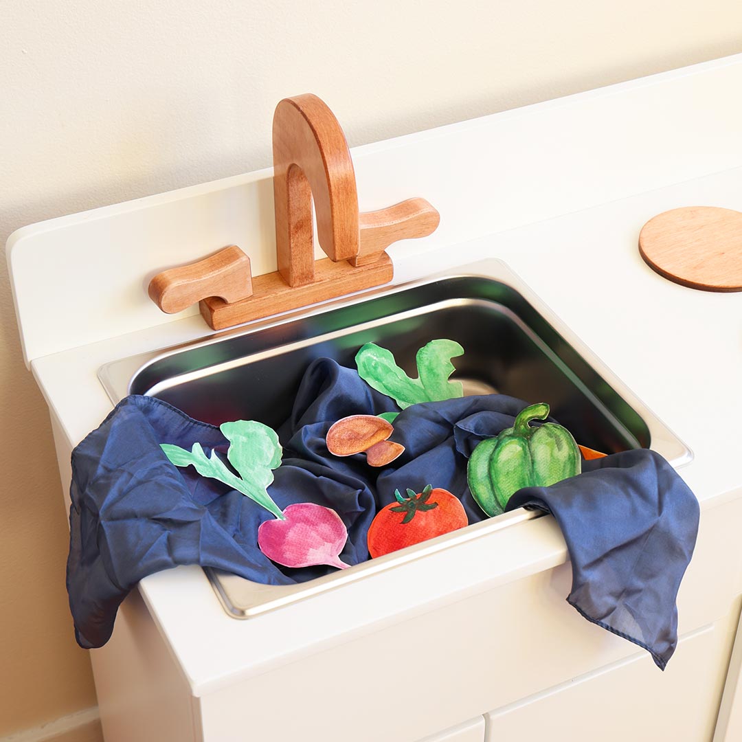 Kitchen sink with a blue play silk draped over the edge, holding paper vegetables.