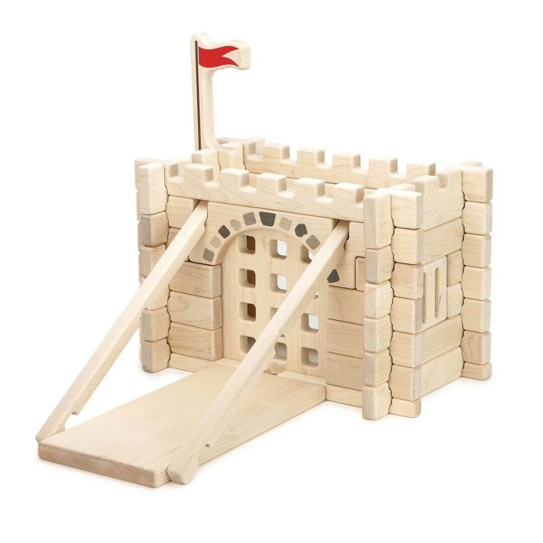 Everwood Friends Wooden Notch Blocks Castle Gate Set assembled into a medieval castle entrance made from natural hard maple wood. The detailed structure features interlocking wooden walls, crenellated battlements, a red flag flying above the castle, and a working portcullis gate beneath a decorative stone-patterned archway. A wooden drawbridge-style ramp extends from the entrance with two angled support beams, showcasing the creative building possibilities of this 47-piece wooden castle set - Bella Luna Toy