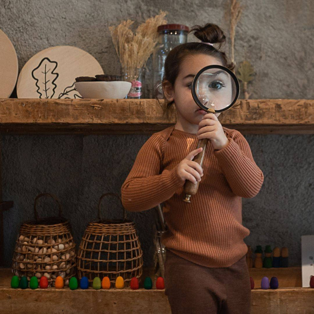 Small child holding a large magnifying glass up to their eye, enlarging it, with Grapat Mandala Wooden Rainbow Eggs lined up on a shelf behind