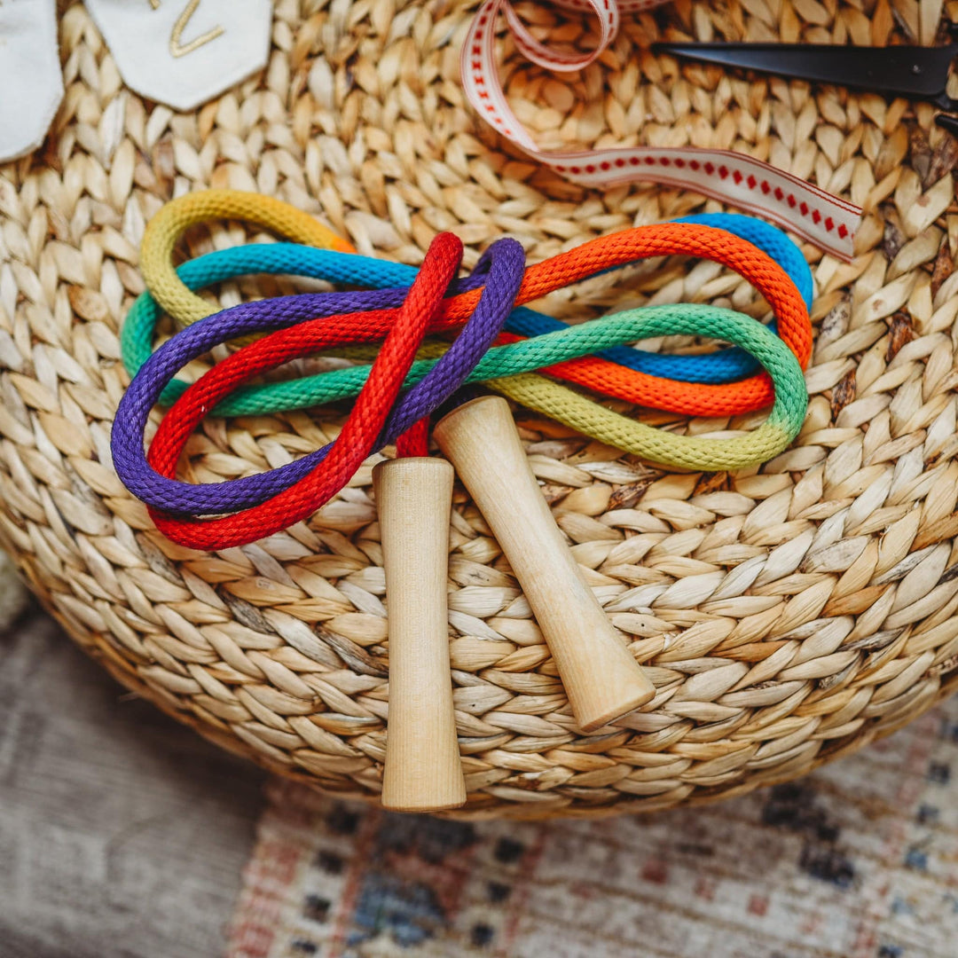 A vibrant, hand-painted Rainbow Jump Rope crafted with smooth wooden handles and sturdy cotton rope set on a woven basket. Bella Luna Toys. 