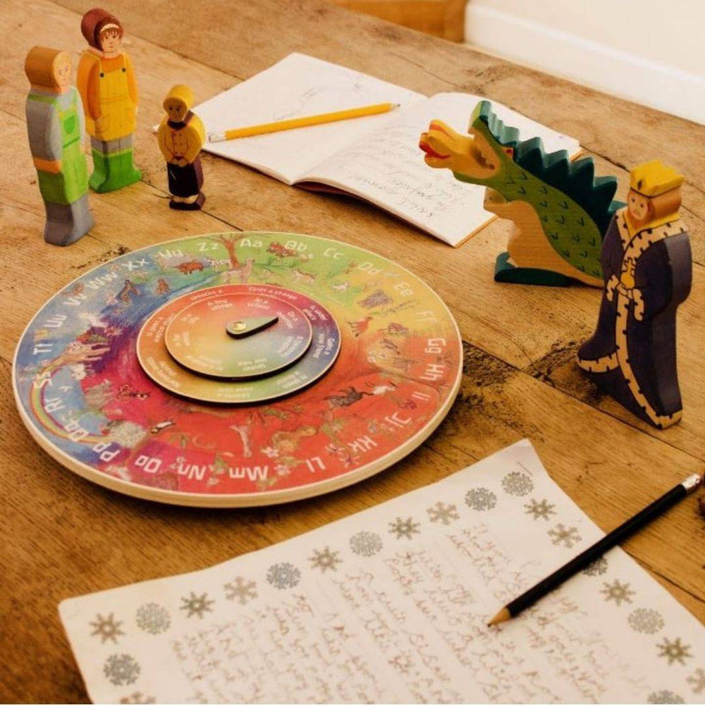 Waldorf Alphabet Letters ABC Wooden Story Wheel on wooden table with notebook, writing paper and pencils, and wooden figures showing dragon, knight, other people