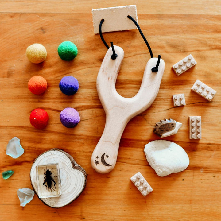 Wooden slingshot lying on wooden table, surrounded by colorful felt ball, brown toy building bricks, clear and green crystals- Bella Luna Toys
