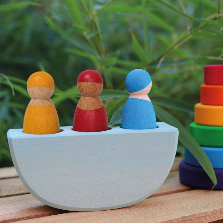 Grimm's 3 in a Wooden Boat shown on a wooden table, alongside a small rainbow stacker, with some greenery in the background