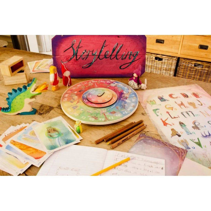 Story Wheel on a wooden table surrounded by some wooden and felt play figures, pencils, notebook, ABC poster
